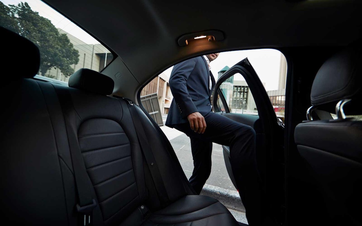 A passenger in a navy blue suit stepping into the backseat of a black luxury sedan at an airport in Chesterfield, Missouri.