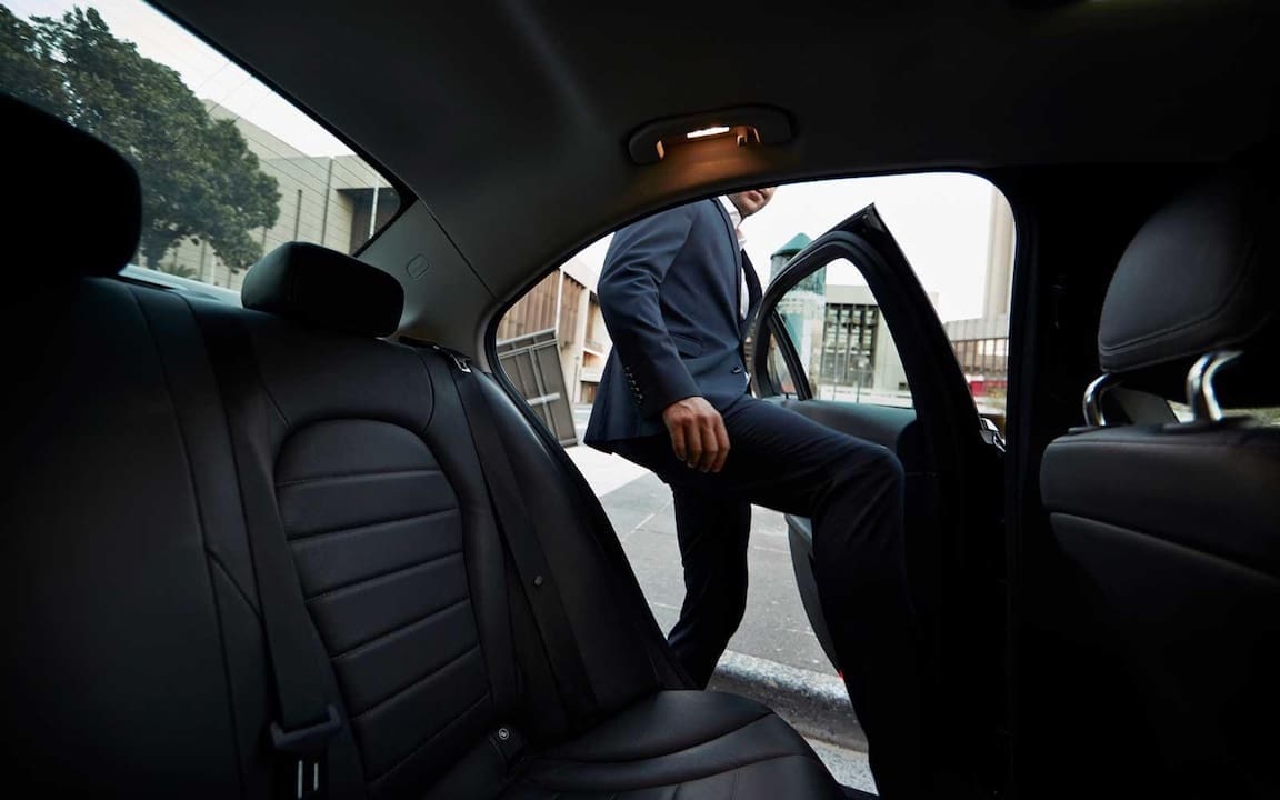 A passenger in a navy blue suit stepping into the backseat of a black luxury sedan at an airport in Chesterfield, Missouri.