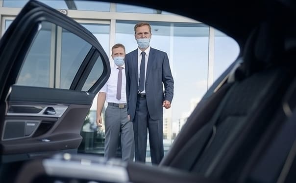 Professional Executive Chauffeur St. Louis A professional chauffeur in a suit and protective face mask holding open the door of a black luxury sedan for an executive client in St. Louis.